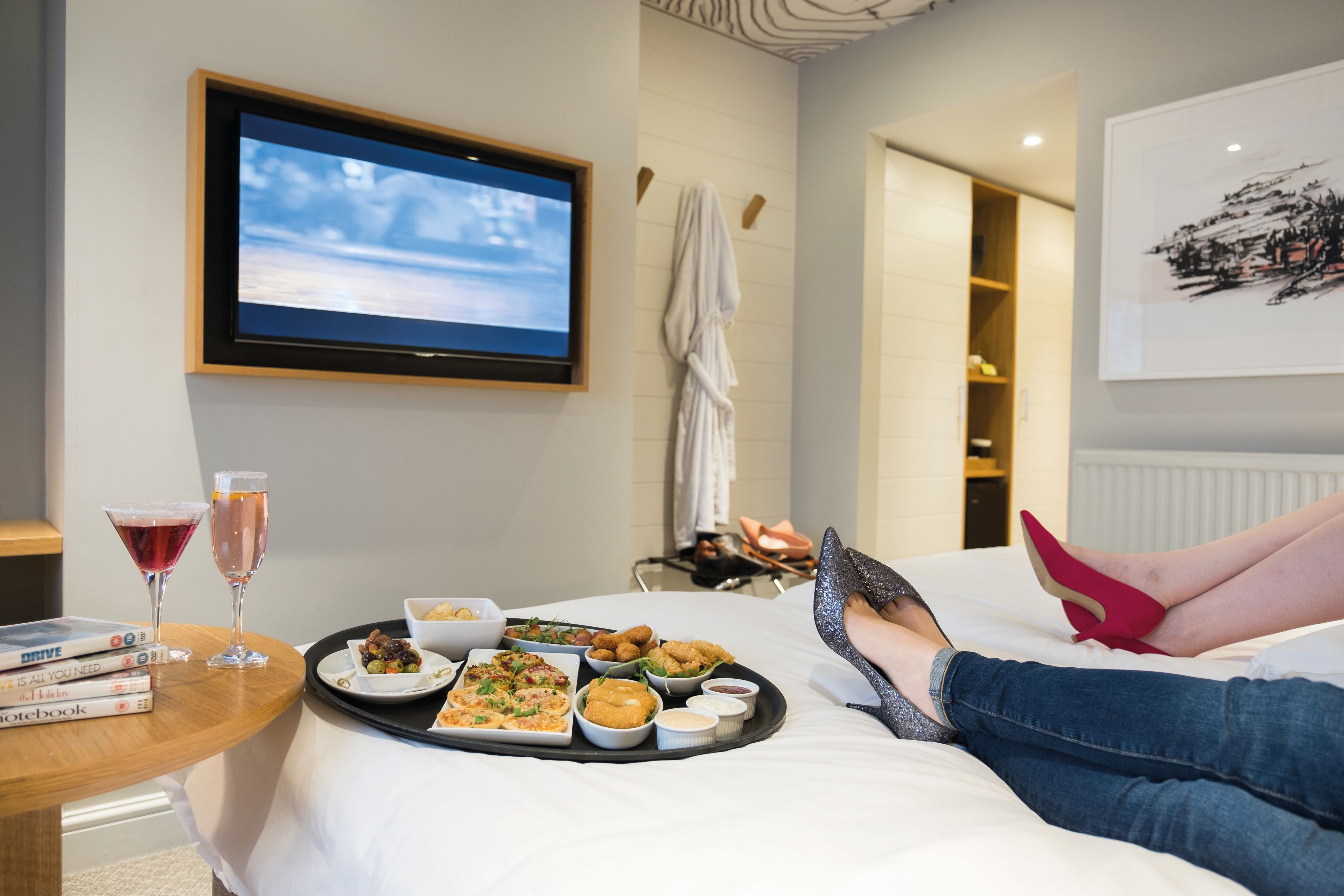 two girls lying on a bed with a tray of food, drinks, and movies