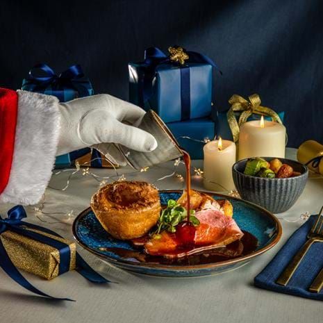 Santa pours gravy on a Christmas dinner plate with a Yorkshire pudding